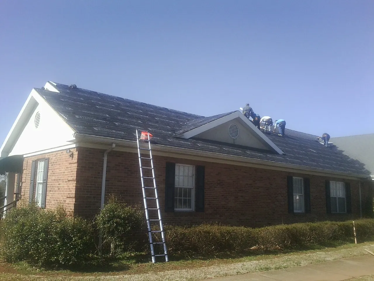 Crew tearing shingles off a brick ranch roof with a ladder set — Emergency Roof Repair work by our crew in Webster